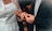 Two individuals participate in a wedding ceremony. The person on the left is dressed in a white gown with delicate lace and sequin details, while the person on the right is wearing a dark suit. They are exchanging rings, symbolizing a marriage commitment.
