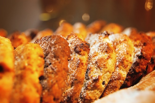 A close-up view of a row of freshly baked cookies, dusted with powdered sugar. The cookies appear golden brown and textured, suggesting they might contain chocolate chips or nuts.