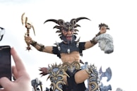 An elaborate and intimidating sculpture depicting a powerful, mythical warrior with horns, holding an axe in one hand and a sculpted head in the other. The warrior is adorned with skulls and intricate armory, displaying a fierce expression. A hand holding a smartphone is visible in the foreground, capturing the scene.