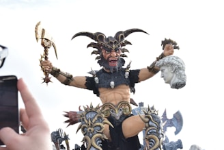 An elaborate and intimidating sculpture depicting a powerful, mythical warrior with horns, holding an axe in one hand and a sculpted head in the other. The warrior is adorned with skulls and intricate armory, displaying a fierce expression. A hand holding a smartphone is visible in the foreground, capturing the scene.