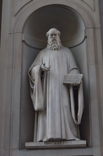 A stone statue of a bearded man in flowing robes stands in an alcove. The figure holds a tablet with one hand while the other is slightly raised in a gesture. The name 'Guido Aretino' is inscribed at the base of the statue.