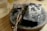 A wooden surface holds a circular metal base on which rests a charred rectangular object alongside a cluster of small, rectangular metal pieces. There is also a pair of tweezers placed on the surface. To the side, a transparent glass holds a light-colored liquid.