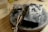 A wooden surface holds a circular metal base on which rests a charred rectangular object alongside a cluster of small, rectangular metal pieces. There is also a pair of tweezers placed on the surface. To the side, a transparent glass holds a light-colored liquid.