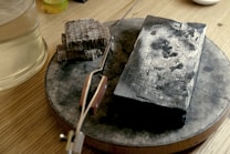 A wooden surface holds a circular metal base on which rests a charred rectangular object alongside a cluster of small, rectangular metal pieces. There is also a pair of tweezers placed on the surface. To the side, a transparent glass holds a light-colored liquid.