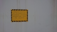 A yellow sign with black diagonal stripes along the border contains text in Greek. The sign is mounted on a light gray, possibly metallic, surface.