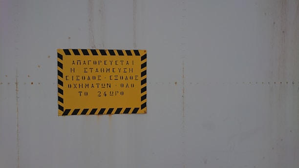 A yellow sign with black diagonal stripes along the border contains text in Greek. The sign is mounted on a light gray, possibly metallic, surface.