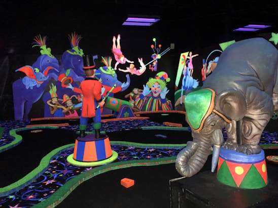 A vibrant, colorful scene reminiscent of a circus with playful elephants, clowns, and acrobats performing. The bright neon colors contrast against a dark backdrop, creating an energetic and whimsical atmosphere. Various performers, including a ringmaster in a red coat, are engaging in entertaining acts, and the scene is filled with detailed, exaggerated circus elements.