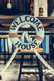 A wooden lifebuoy with the words 'Welcome House' painted on it is hanging from a rope. The lifebuoy is mounted on a wooden structure, with bar stools visible in the background. The setting appears to be nautical-themed, suggesting a cozy and inviting atmosphere.