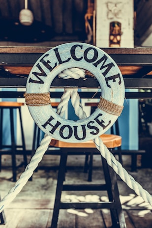 A wooden lifebuoy with the words 'Welcome House' painted on it is hanging from a rope. The lifebuoy is mounted on a wooden structure, with bar stools visible in the background. The setting appears to be nautical-themed, suggesting a cozy and inviting atmosphere.