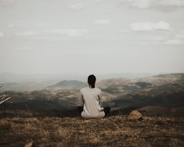 A photo of a woman sitting on the top of a mountain, looking out at the view. We see her from behind. It's peaceful.