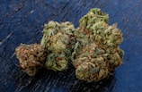 A close-up view of cannabis buds with a dense and frosty appearance, displaying various shades of green with hints of orange and brown hairs, resting on a dark, textured surface.