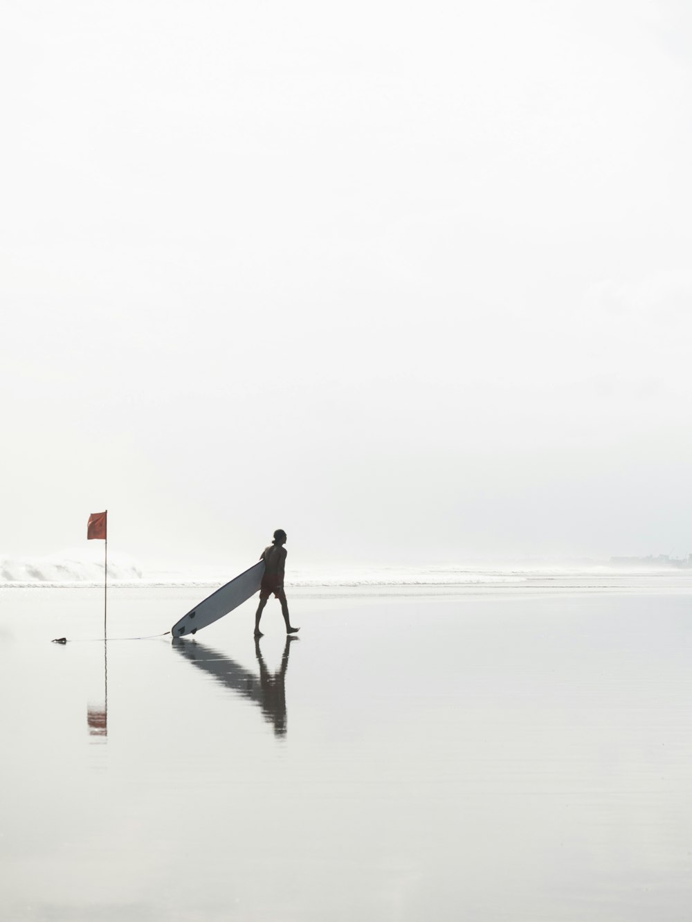 日中、旗の近くのビーチでサーフボードを引きずりながら歩く男