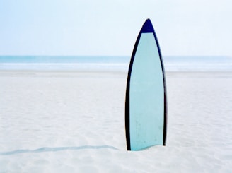 A serene shot of a surfboard standing upright in the shallow water, waves gently rolling in the background.