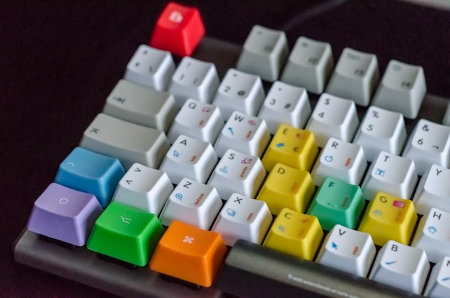 A mechanical keyboard with a variety of colorful keycaps, including red, blue, green, yellow, and purple. The keys contain a mix of English letters, symbols, and playful icons. The background is dark, making the colors of the keys stand out vividly.
