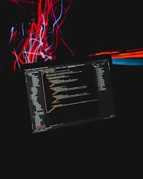A computer screen displaying code is set against a dark background with dynamic streaks of red and blue light. The code appears to be in a text editor or IDE, with a clear folder structure on the left and lines of syntax-highlighted text taking up the central portion. The colorful light streaks add a sense of motion and energy.