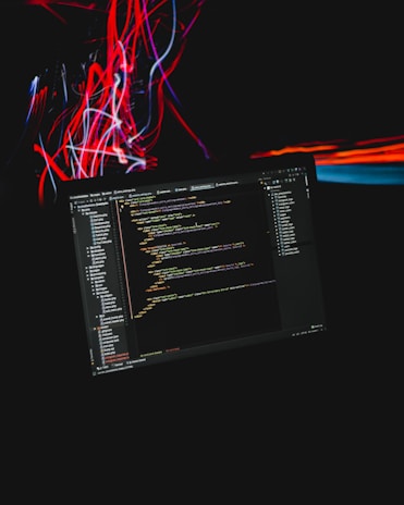 A computer screen displaying code is set against a dark background with dynamic streaks of red and blue light. The code appears to be in a text editor or IDE, with a clear folder structure on the left and lines of syntax-highlighted text taking up the central portion. The colorful light streaks add a sense of motion and energy.