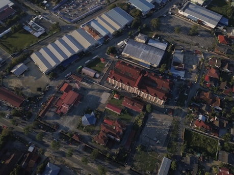 An aerial view of an urban area featuring several large warehouse buildings with light-colored roofs, surrounded by roads and smaller structures. The central part of the image shows a complex of red-roofed buildings, possibly residential or commercial, with several trees and greenery scattered throughout the area. Paved and unpaved surfaces are visible, with some parked vehicles.