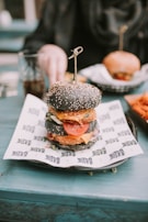 A gourmet burger with a black sesame seed bun is placed on a piece of printed paper atop a plate. Inside the burger, layers of fresh lettuce, tomato slices, cheese, and a breaded patty are visible. In the background, a hand reaches for a drink on the teal table, and another burger is slightly out of focus.