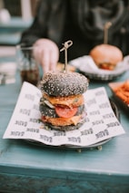 A gourmet burger with a black sesame seed bun is placed on a piece of printed paper atop a plate. Inside the burger, layers of fresh lettuce, tomato slices, cheese, and a breaded patty are visible. In the background, a hand reaches for a drink on the teal table, and another burger is slightly out of focus.