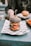 A gourmet burger with a black sesame seed bun is placed on a piece of printed paper atop a plate. Inside the burger, layers of fresh lettuce, tomato slices, cheese, and a breaded patty are visible. In the background, a hand reaches for a drink on the teal table, and another burger is slightly out of focus.