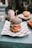 A gourmet burger with a black sesame seed bun is placed on a piece of printed paper atop a plate. Inside the burger, layers of fresh lettuce, tomato slices, cheese, and a breaded patty are visible. In the background, a hand reaches for a drink on the teal table, and another burger is slightly out of focus.