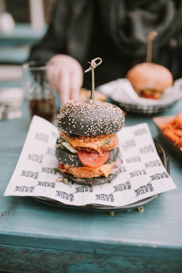 A gourmet burger with a black sesame seed bun is placed on a piece of printed paper atop a plate. Inside the burger, layers of fresh lettuce, tomato slices, cheese, and a breaded patty are visible. In the background, a hand reaches for a drink on the teal table, and another burger is slightly out of focus.