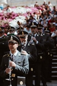A group of uniformed individuals is playing clarinets while standing on stairs. They are wearing formal military-style uniforms and hats, suggesting a ceremonial or formal event. A large crowd is seated behind them, with vibrant pink and white flowers in the background.