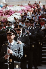 A group of uniformed individuals is playing clarinets while standing on stairs. They are wearing formal military-style uniforms and hats, suggesting a ceremonial or formal event. A large crowd is seated behind them, with vibrant pink and white flowers in the background.