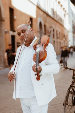 A man dressed in a white suit and shirt plays a violin in a narrow street. The street has a European ambiance, with old, beige-colored buildings lining the sides. A bicycle is visible to the right, and a few people walk in the background.