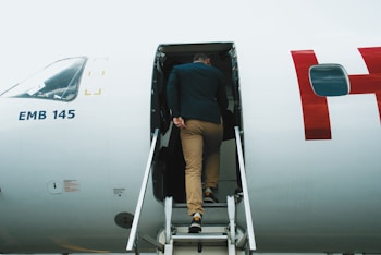 A person wearing a black jacket and beige pants is seen boarding an aircraft via a stairway attached to the plane. The aircraft is marked with the model number 'EMB 145' and features a white exterior with a partial red letter visible.
