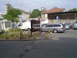 A parking area with several vehicles including a van and a silver station wagon. There's a large sign with Cyrillic letters, a small green patch with grass and weeds, and bollards with black and yellow stripes. Buildings with various architectural styles are visible in the background, including one with a domed roof and a cross.
