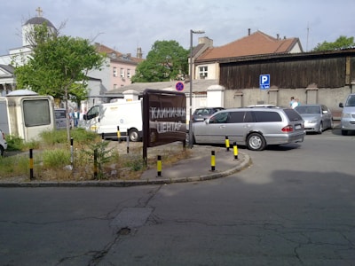 A parking area with several vehicles including a van and a silver station wagon. There's a large sign with Cyrillic letters, a small green patch with grass and weeds, and bollards with black and yellow stripes. Buildings with various architectural styles are visible in the background, including one with a domed roof and a cross.