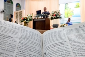 An open book displaying text in focus, likely a Bible, with a blurred background showing a church or religious setting. A speaker stands at a lectern adorned with floral arrangements, and a person is seated nearby.