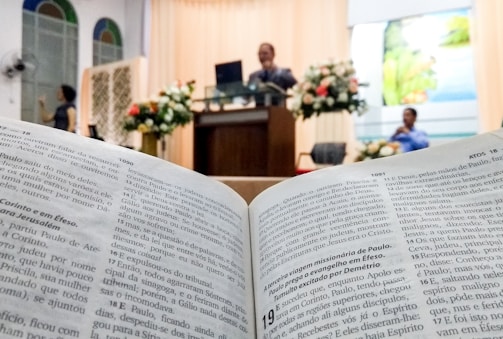 An open book displaying text in focus, likely a Bible, with a blurred background showing a church or religious setting. A speaker stands at a lectern adorned with floral arrangements, and a person is seated nearby.