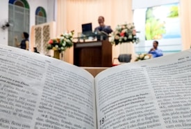 An open book displaying text in focus, likely a Bible, with a blurred background showing a church or religious setting. A speaker stands at a lectern adorned with floral arrangements, and a person is seated nearby.