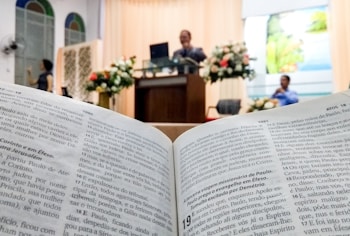 An open book displaying text in focus, likely a Bible, with a blurred background showing a church or religious setting. A speaker stands at a lectern adorned with floral arrangements, and a person is seated nearby.