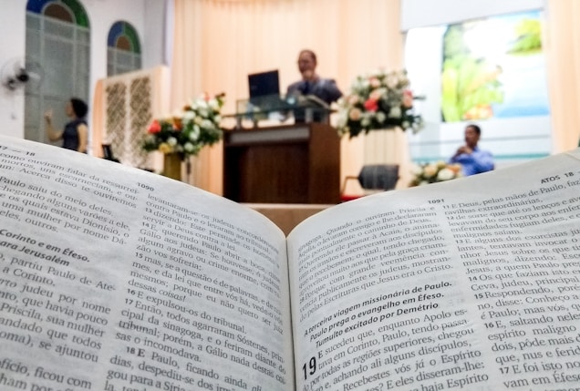 An open book displaying text in focus, likely a Bible, with a blurred background showing a church or religious setting. A speaker stands at a lectern adorned with floral arrangements, and a person is seated nearby.
