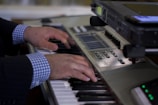 Hands are playing on a musical keyboard with a checkered shirt and a suit jacket visible. The scene includes a high-tech keyboard with numerous control buttons and a screen, possibly for digital sound mixing or music production.