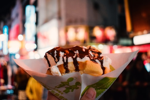 A hand holding a waffle topped with whipped cream and drizzled with chocolate sauce, wrapped in patterned paper. The background features a bustling street scene with bright lights and blurred buildings, suggesting a vibrant urban nightlife setting.