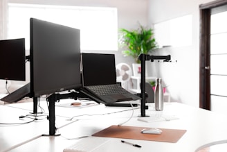 A modern office workspace featuring multiple computer monitors and a laptop on adjustable arms, positioned on a white desk. The desk is equipped with a keyboard, mouse, and a silver water bottle. In the background, there is a green potted plant and a fan, with ample natural light coming through large windows.