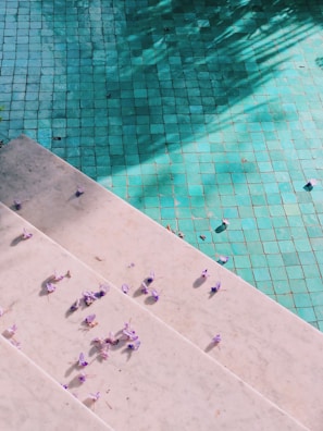 A tiled swimming pool with a light greenish-blue hue is partially covered by shadows of nearby trees. A set of white stone steps descends into the pool, scattered with small purple flowers, adding a delicate touch to the scene.