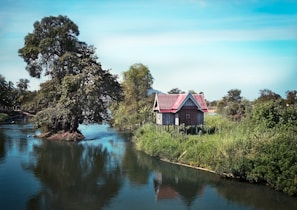 A wooden house with a red roof stands on the edge of a river, surrounded by lush greenery and trees. The water reflects the vibrant colors of the sky and landscape, creating a serene and picturesque setting.