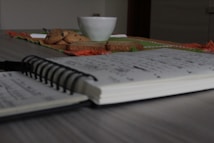 A notebook with handwritten notes lies open on a wooden table. In the background, a white cup, cookies, and slices of bread are arranged on a colorful tablecloth.