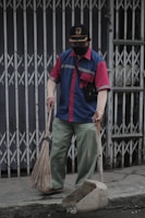 A person wearing a blue and red uniform, a cap, and a face mask holds a broom and a dustpan. They are standing on a sidewalk in front of a closed metal gate, appearing to be cleaning the area.