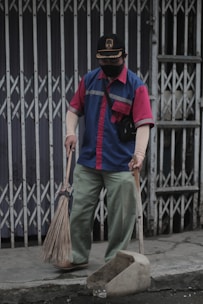 A person wearing a blue and red uniform, a cap, and a face mask holds a broom and a dustpan. They are standing on a sidewalk in front of a closed metal gate, appearing to be cleaning the area.