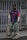 A person wearing a blue and red uniform, a cap, and a face mask holds a broom and a dustpan. They are standing on a sidewalk in front of a closed metal gate, appearing to be cleaning the area.