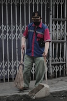 A person wearing a blue and red uniform, a cap, and a face mask holds a broom and a dustpan. They are standing on a sidewalk in front of a closed metal gate, appearing to be cleaning the area.