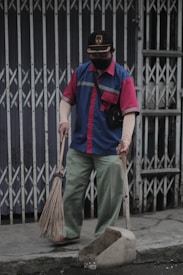 A person wearing a blue and red uniform, a cap, and a face mask holds a broom and a dustpan. They are standing on a sidewalk in front of a closed metal gate, appearing to be cleaning the area.