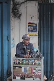 A man in a cap and jacket stands behind a glass counter filled with various watches and boxes. The background features a white wall with several posters and a blue metal structure on the left. A small black bag is placed on top of the counter, and there are signs above advertising watch repair services.