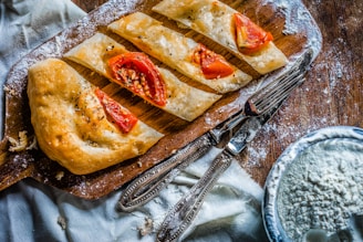 A wooden cutting board displays slices of baked bread adorned with tomato segments and herbs. The surface is dusted with flour, suggesting a baking or cooking activity. Beside the board, there are utensils and a bowl containing a powdery substance, likely more flour, on a rustic wooden table.